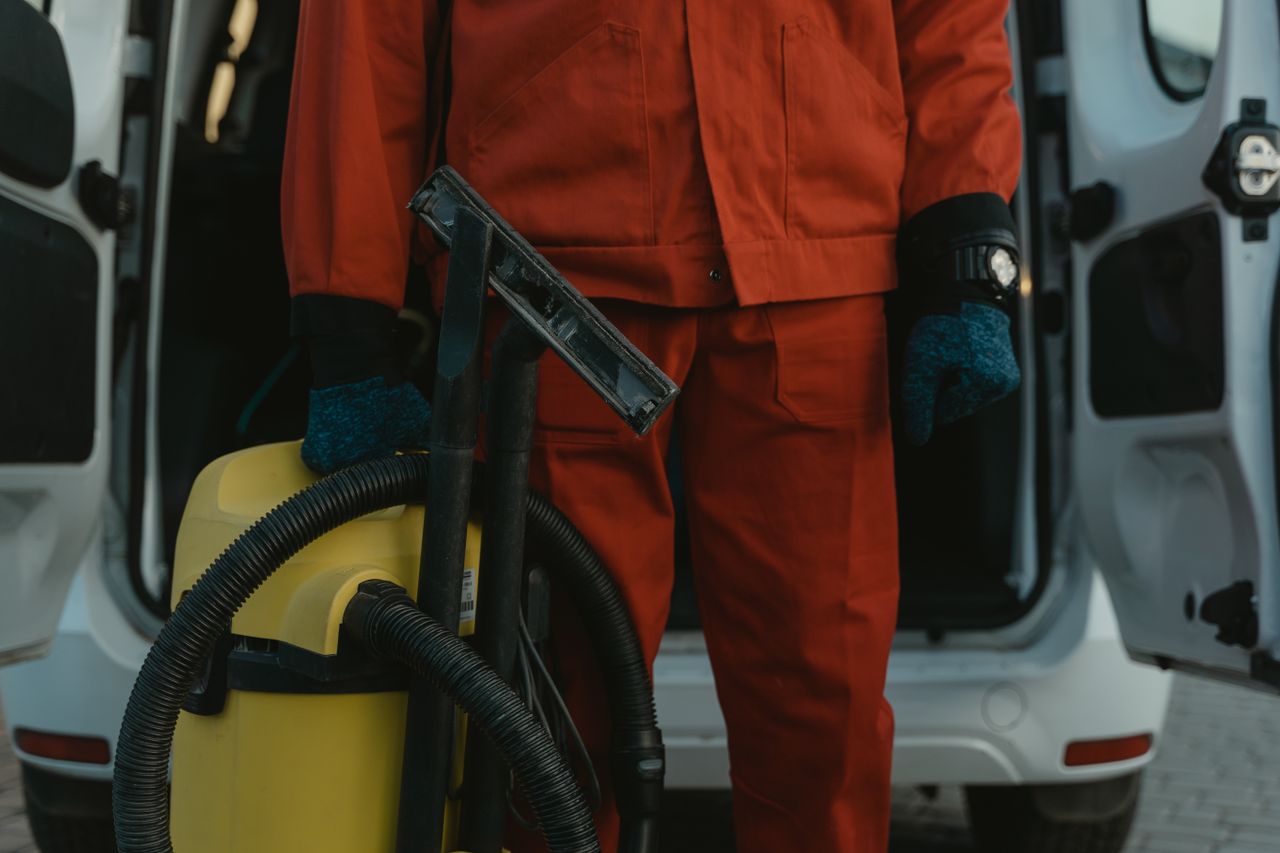 services-03 Un professionnel du nettoyage en uniforme rouge tient un aspirateur près d’une camionnette, prêt à intervenir.